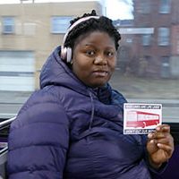 woman on bus in puffy coat and headphone holding a postcard that says bus lines are life lines
