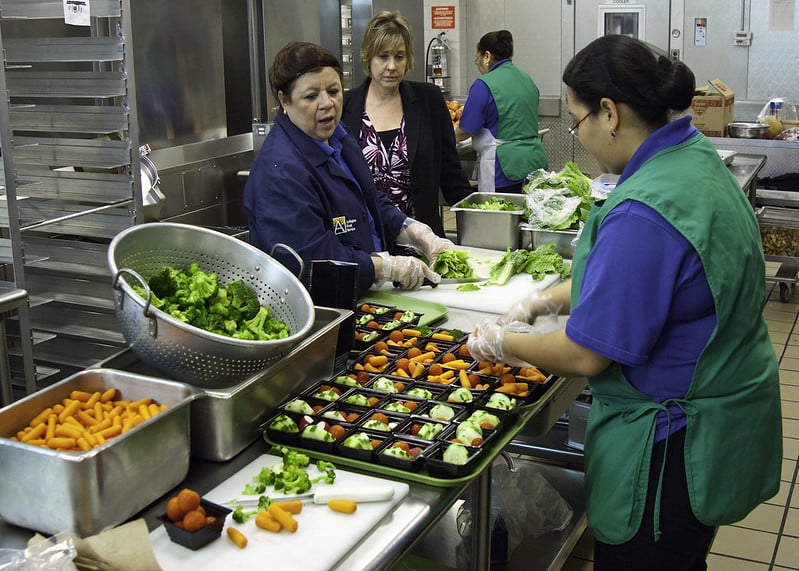 A cafeteria manager and worker, wearing uniforms, hairnets and plastic gloves, prep small servings of salad in an industrial kitchen while the district nutrition director looks on, all women. (USDA/flickr)